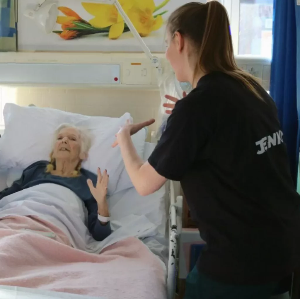 Patients in hospital beds participating in a seated dance session led by an instructor