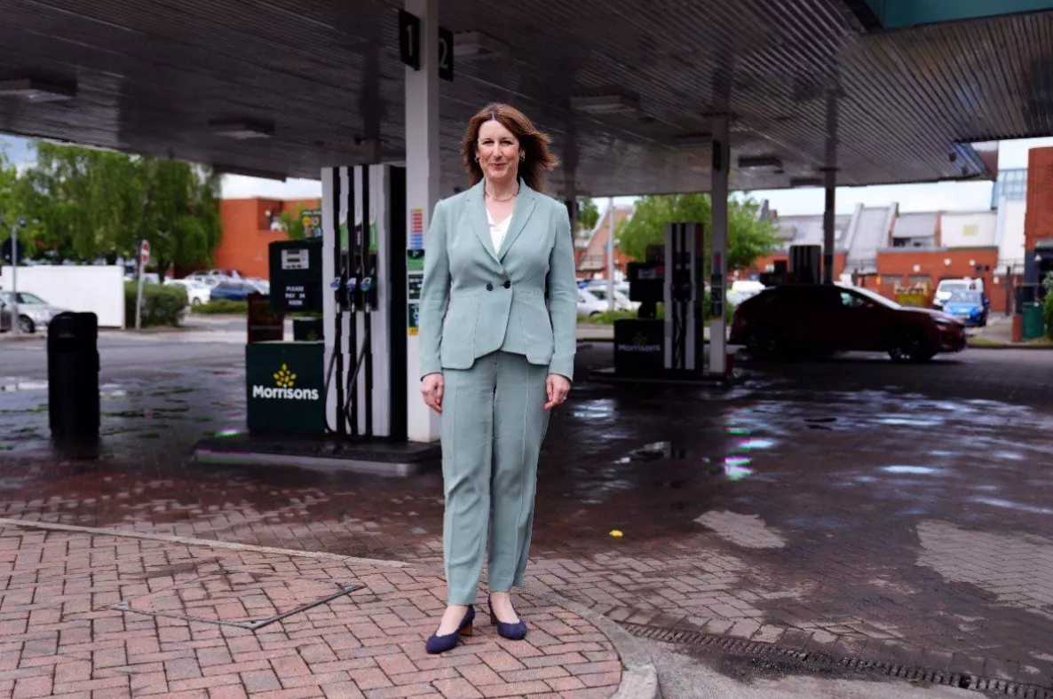 A young woman walking past a petrol station with a worried expression, clutching a shopping bag.